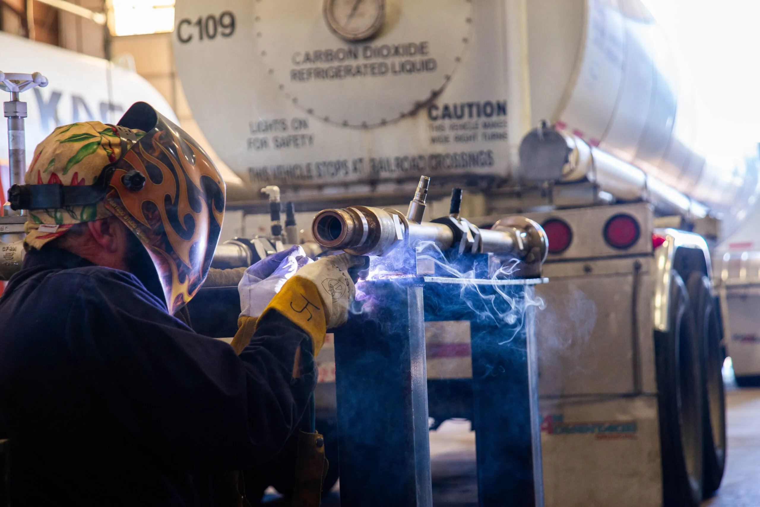 Welder in a flame-decorated helmet welding a pipe in a workshop with a carbon dioxide refrigerated liquid tanker in the background