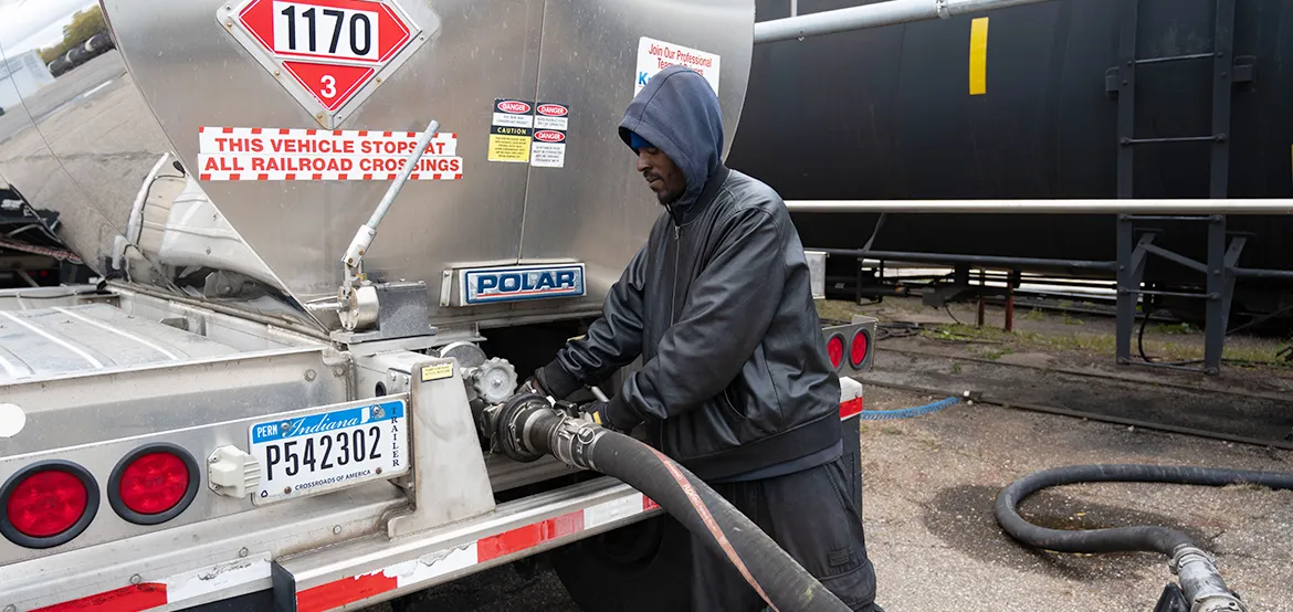 Worker connecting a transfer hose to the rear valve of a tanker trailer at a transloading site near a rail car