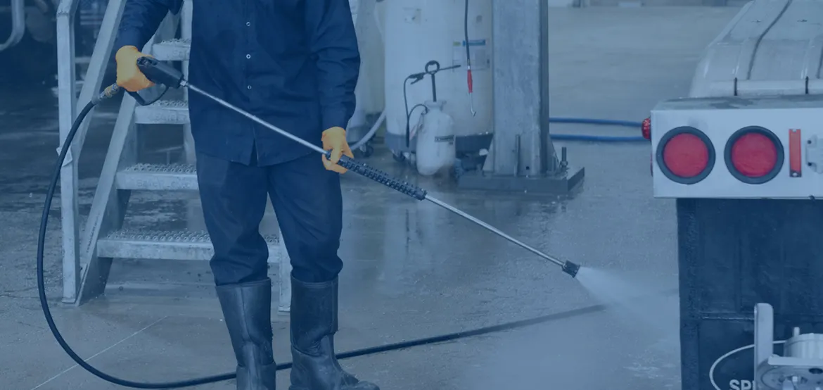 Worker in safety gear pressure-washing the ground near a tanker truck at a facility