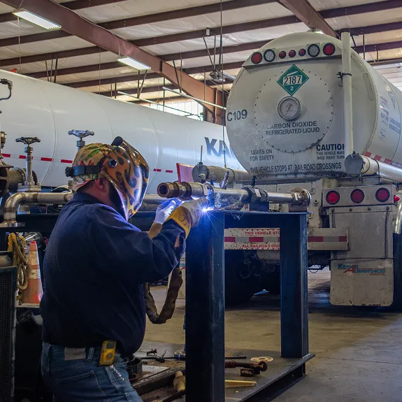 Welder in protective gear working in a maintenance shop with a KAG carbon dioxide tanker in the background