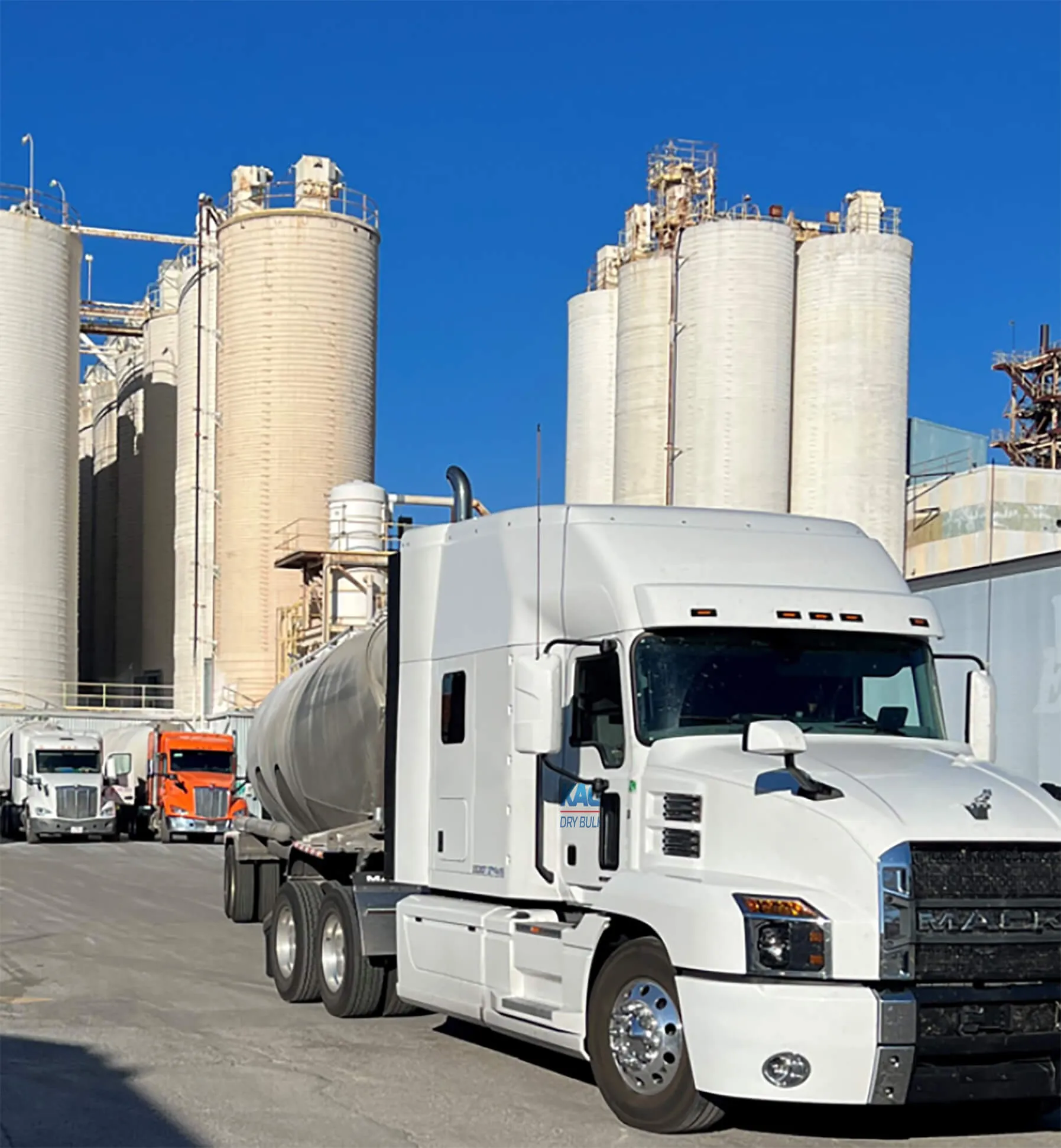 Dry bulk tanker truck parked in front of large industrial storage silos under a bright blue sky